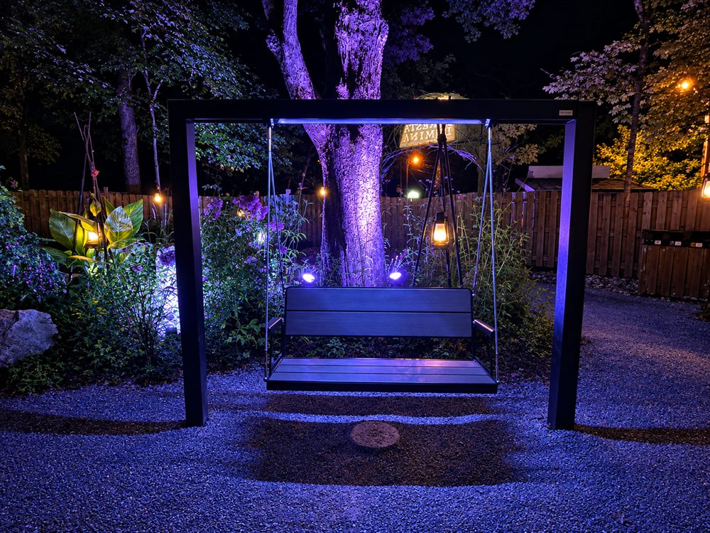 Foresta Lumina, à Coaticook : la balançoire suspendue sous son portique d'acier noir, au-dessus du gravier du sentier, éclairée de lanternes et de lumière violette au pied d'un grand arbre.