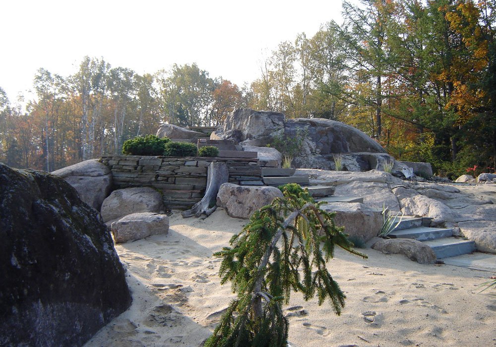 Escalier de pierre et mur de pierre sèche posés entre de grands blocs erratiques, sur un lit de sable pâle, avec un conifère pleureur et des touffes de yucca, devant la forêt d'automne.