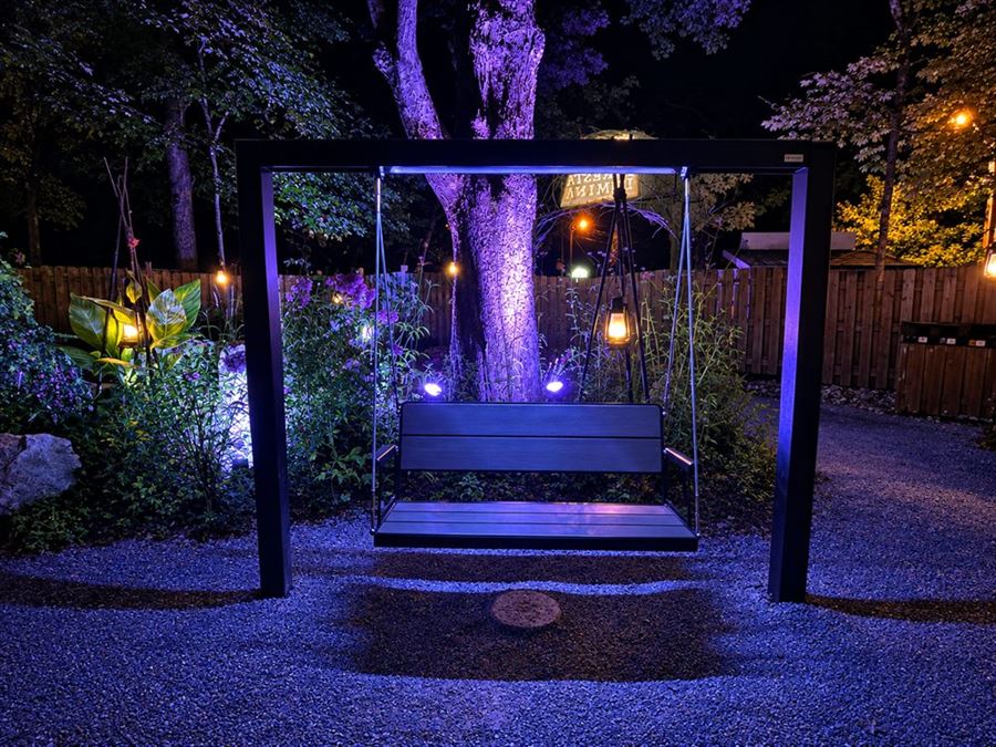 Foresta Lumina, à Coaticook : la balançoire suspendue sous son portique d'acier noir, au-dessus du gravier du sentier, éclairée de lanternes et de lumière violette au pied d'un grand arbre.