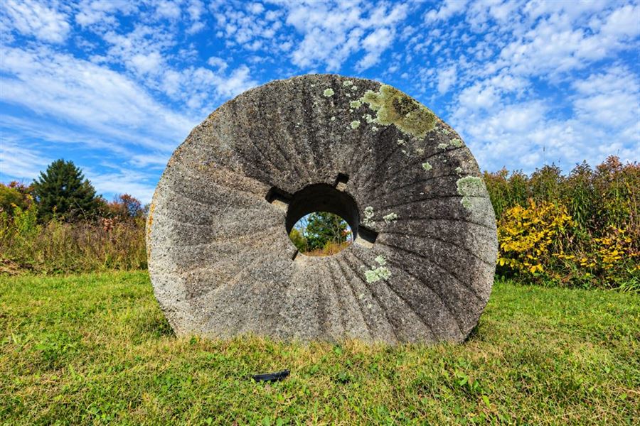Meule de moulin en pierre dressée dans l'herbe du jardin de Sutton : les raies rayonnantes taillées dans le granite, le lichen vert et le trou carré du centre, sous un ciel bleu semé de nuages.
