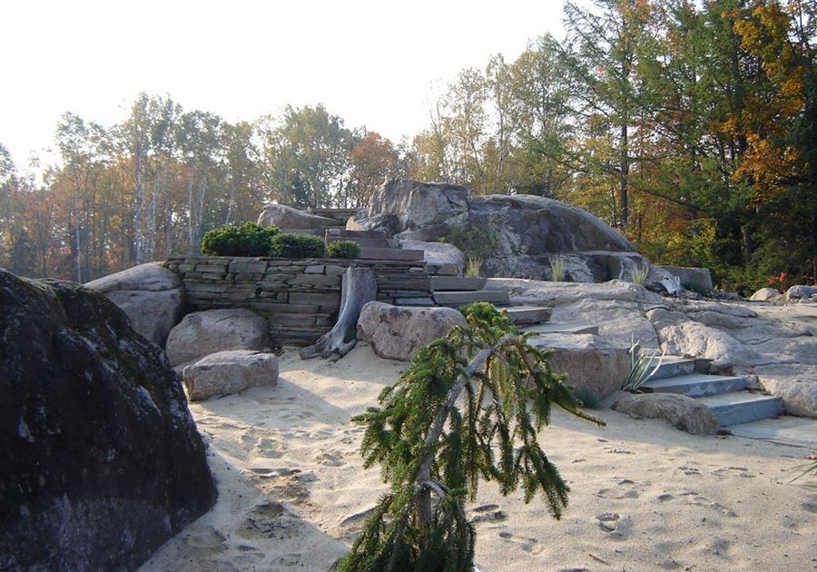 Escalier de pierre et mur de pierre sèche posés entre de grands blocs erratiques, sur un lit de sable pâle, avec un conifère pleureur et des touffes de yucca, devant la forêt d'automne.